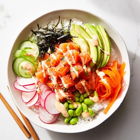 A chef's spoon drizzling spicy mayo over a colorful Poke Bowl with edamame and nori strips on a marble surface.